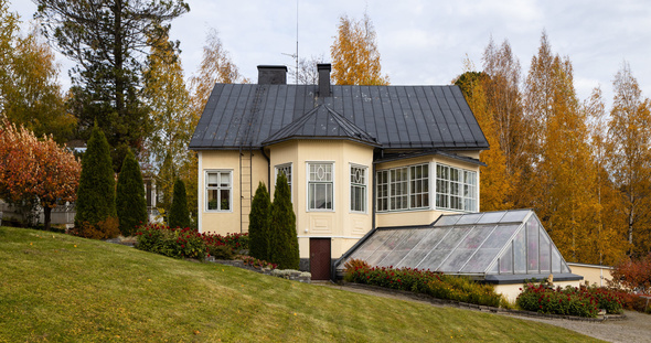 Keltainen rakennus syksyisessä maisemassa. Kuvan etualalla on hirveä nurmikko. Rakennuksen vieressä ja takana on erilaisia puita. Rakennuksen edustalla on kasvihuone.