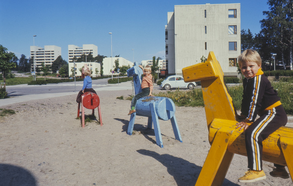 Lapset ratsastavat puuhevosilla kerrostaloalueen leikkipihalla 1970-luvulla