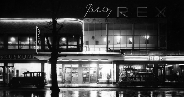 Matalan rakennuksen ikkunoista loistaa valoa pimeään ja sateiseen katukuvaan. Rakennuksen katolla on valomainos, jossa lukee "Bio Rex". Rakennuksen eteen on pysäköity muutamia autoja.