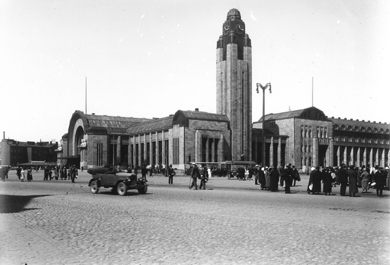 Korkean tornirakennuksen edustalla vilisee ihmisiä ja kulkuneuvoja. Kuvan edustalla näkyy mukulakivetys.