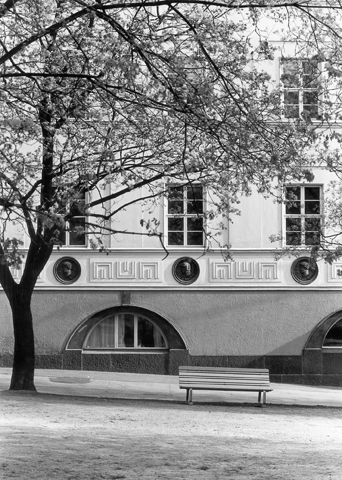 Valokuva Helsingin ruotsalaisen tyttökoulun julkisivusta. Julkisivussa klassisia koristeaiheita ja rakennuksen edustalla kasvaa puu jonka vieressä on penkki.