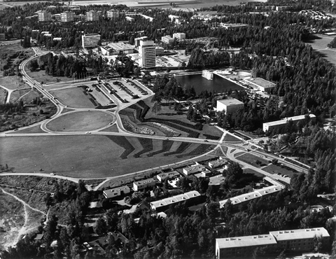 Ilmakuva Tapiolan keskustasta. Edustalla kerros- ja rivitaloja, sen takana teitä ja niittyaukeaa ja näiden takana Tapiolan keskusallas torneineen.