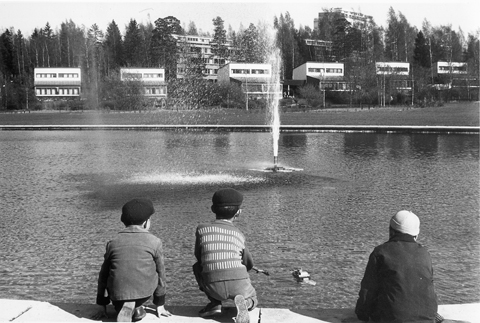 Lapsia Tapiolan keskustassa, vesialtaan edustalla. Kuvassa etualalla kolme poikaa selin kuvaajaan päin, taaempana vesiallas suihkulähteineen, jonka takana pienkerrostaloja ja korkeampia kerrostaloja.
