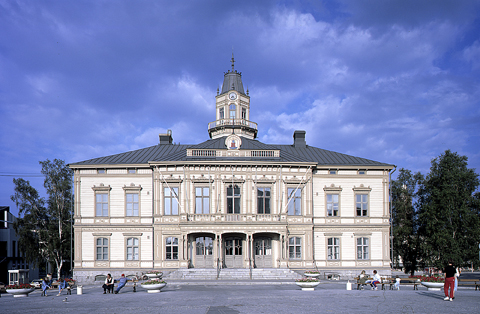 Eskteriöörivalokuva Pietarsaaren raatihuoneesta. Valokuva on otettu suoraan rakennuksen edustalta, puinen rakennus sijaitsee torin laidalla, on kaksikerroksinen ja tyyliltään uusrenessanssinen.