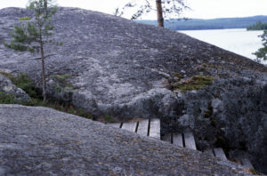 Puiset portaat upotettuina luonnon kallioon. Taustalla näkyy muutama puu ja järvimaisema.