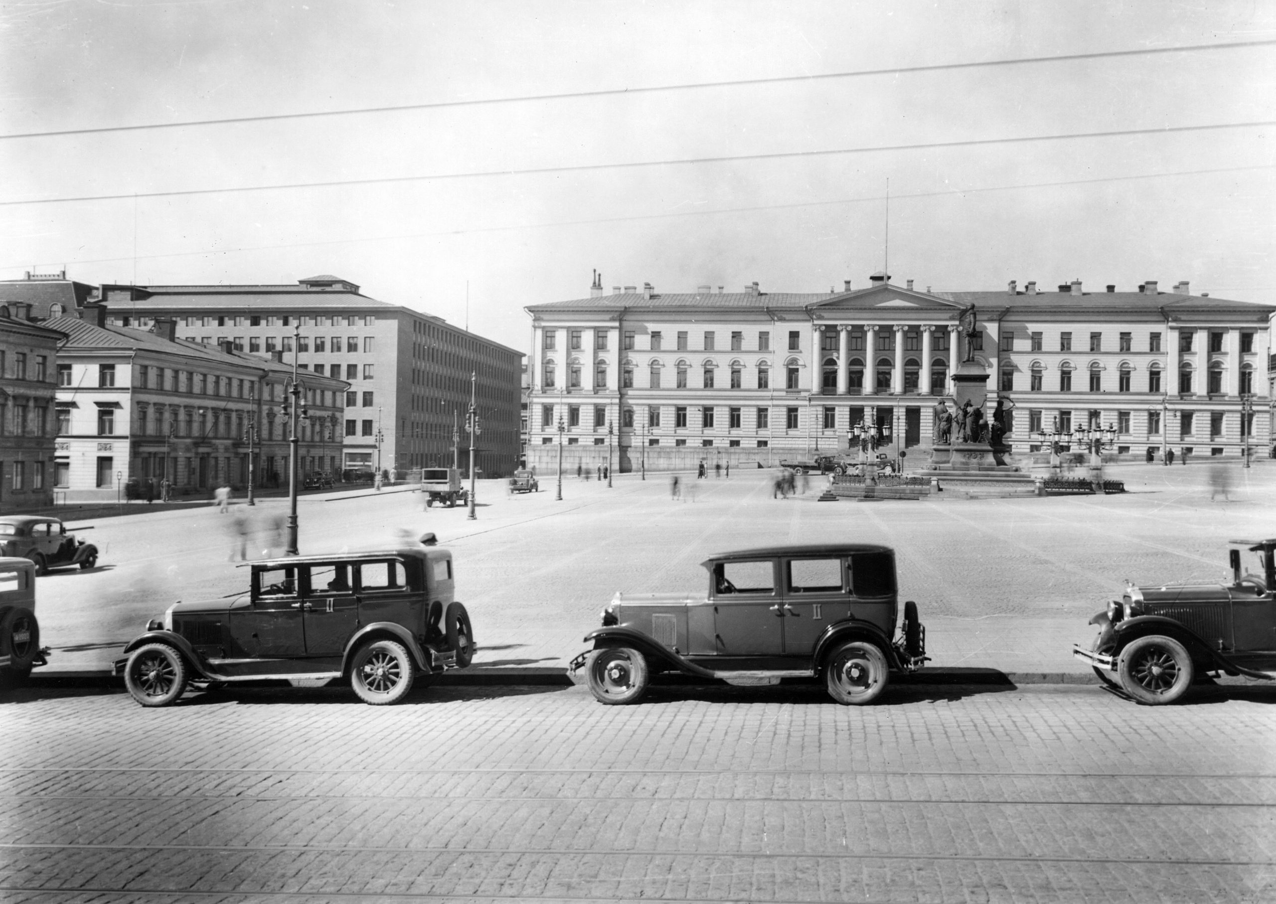 Mustat autot pysäköitynä aution senaatintorin reunalla. Taustalla näkyy vanha Helsingin yliopistorakennus.