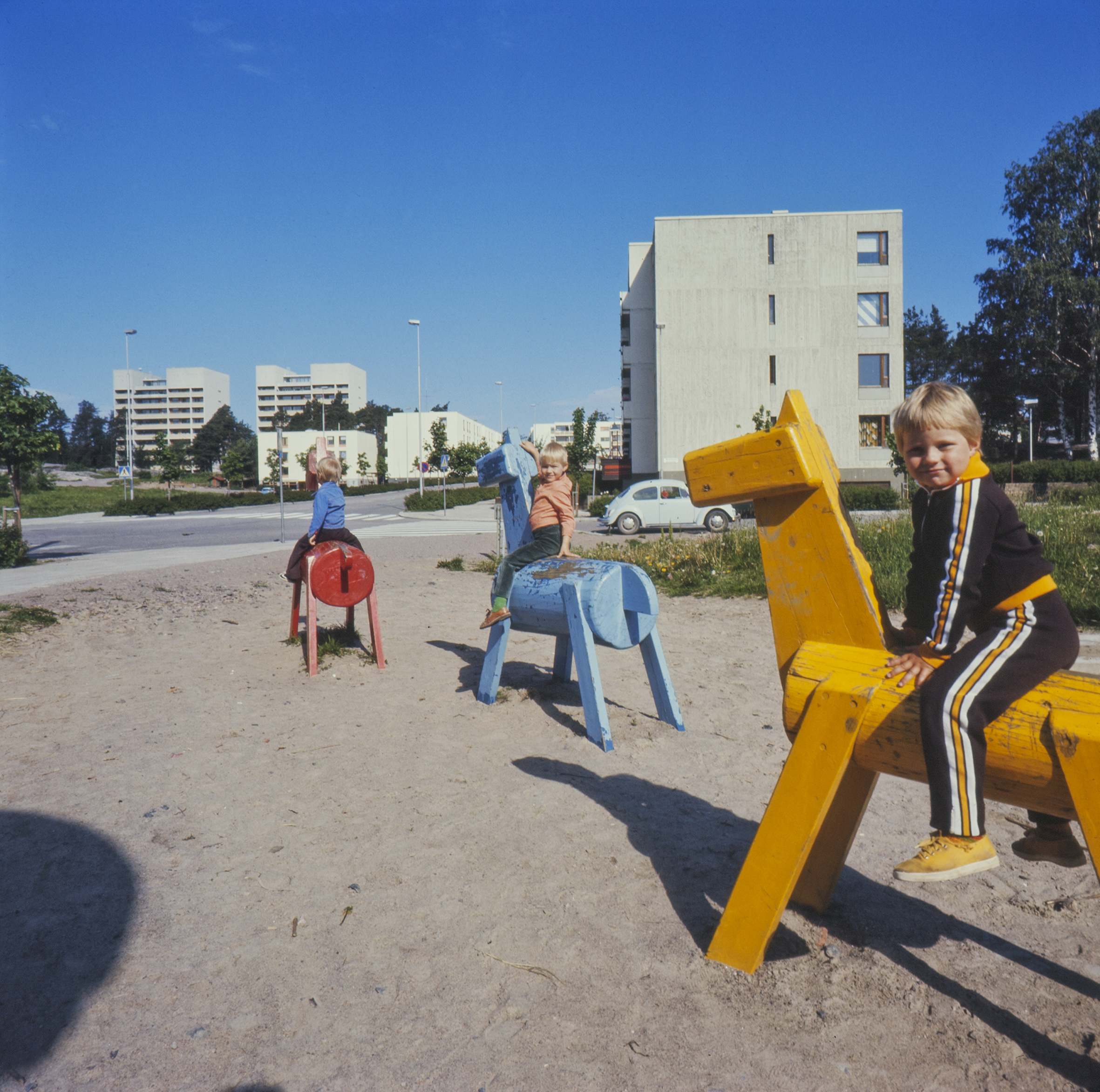 Lapset ratsastavat puuhevosilla kerrostaloalueen leikkipihalla 1970-luvulla